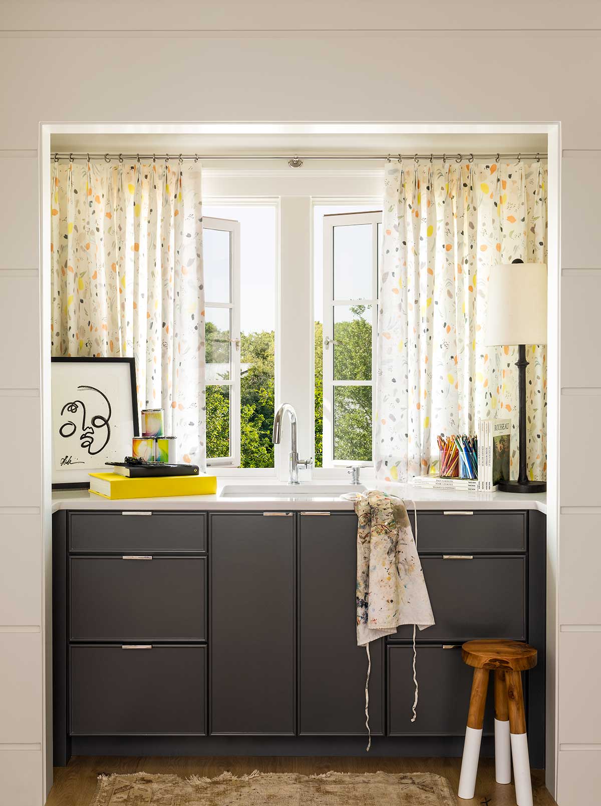 A sink with Marvin Ultimate Casement Push Out windows and colorful window treatments, an artist's apron, and art supplies.